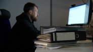 Ein Mann sitzt vor einem Computer, im Vordergrund liegt ein Aktenstapel © NDR 