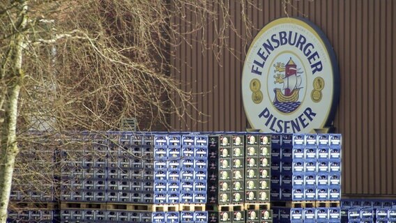 Zahlreiche Kisten stehen gestapelt auf Paletten auf dem Gelände der Brauerei in Flensburg vor dem großen Firmenlogo. © NDR 