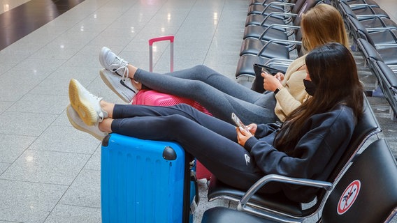 Zwei Frauen sitzen in einer Wartehalle eines Flughafens. Sie haben ihre Beine auf ihren Koffern abgelegt. © Imago Foto: Rupert Oberhäuser