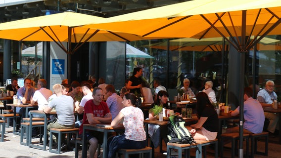 Mehrere Menschen sitzen bei guten Wetter unter den gelben Sonnenschirmen eines Cafés. © NDR Foto: Anna-Lena Trey