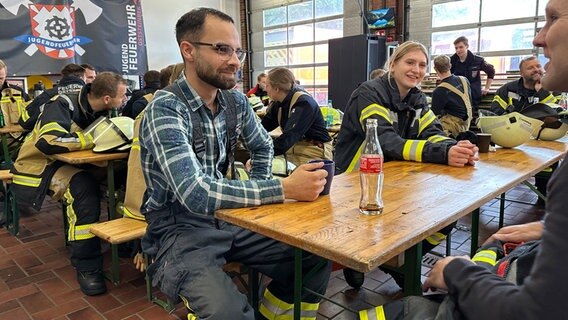 Ein Mann sitzt mit den Einsatzkräften einer Feuerwehr in deren Halle. © NDR Foto: Johannes Tran