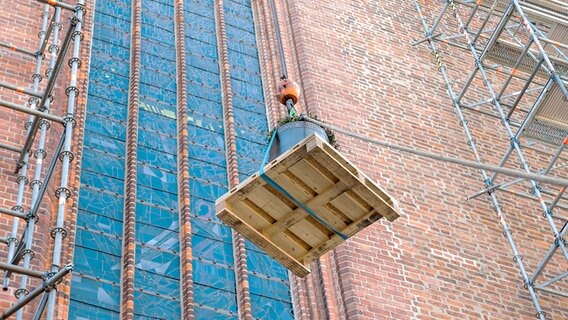 Eine Glücke auf einer Palette befestigt wird an der Außenfassade der Lübecker Marienkirche hochgezogen. Daneben befinden sich Gerüste. © dpa-Bildfunk Foto: Daniel Bockwoldt/dpa-Bildfunk
