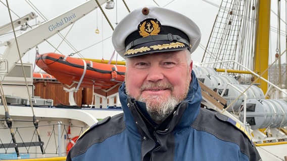 Der Marinekapitän des Schiffs lächelt in Uniform in die Kamera. Im Hintergrund sind Taue, Segel und Masten an Deck der Gorch Fock zu sehen.  Foto: Christian Wolf