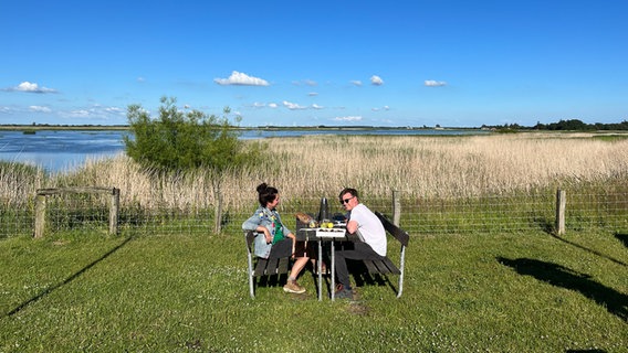 Zwei Menschen sitzen auf einer Wiese an der Küste an einem Tisch. © NDR Foto: Kai Küken