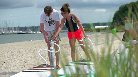 Zwei Menschen pumpen ihre SUP-Boards am Strand auf. © NDR Foto: Lena Storm