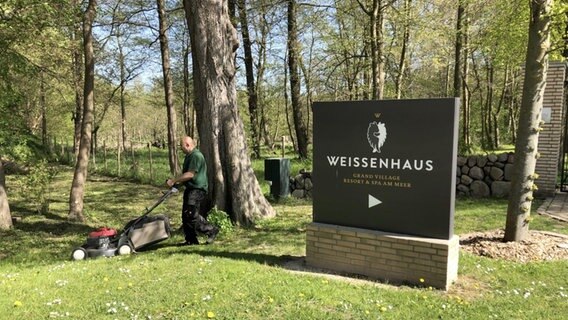 Ein Mann mäht Gras auf einer Wiese vor einem Hotel-Ressort. © NDR Foto: Hauke von Hallern