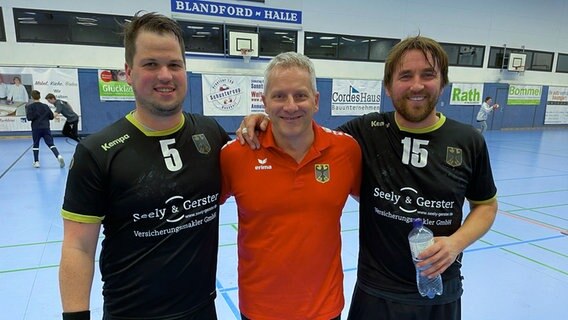 Drei Männer lächeln in die Kamera während eines Handball-Spiels. Drei Männer lächeln in die Kamera während eines Handball-Spiels. Foto: Finn-Ole Martins