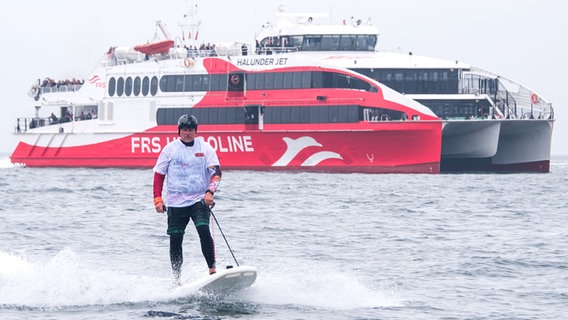 Jörg Singer, Bürgermeister der Insel Helgoland, fährt im Rahmen einer Aktion gegen Plastikmüll im Meer auf einem E-Surfboard neben dem Katamaran «Halunder Jet». © dpa Foto: Daniel Bockwoldt