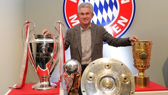 Jupp Heynckes posiert mit seinen 2013 gewonnenen Pokalen vor dem Logo des FC Bayern München. © picture alliance Foto: Frank Hoermann/SVEN SIMON