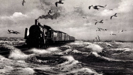 Ein Postkartenfoto aus den dreißiger Jahren des Hindenburgdamms, auf dem eine Dampflok fährt. Das Wasser schlägt hohe Wellen und es fliegen einige Möwen um die Lok herum. © imago/Arkivi 