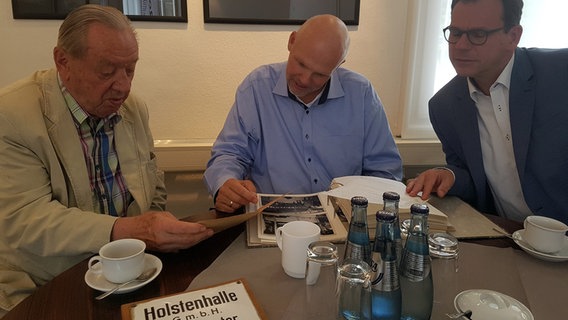 Herbert Möller (l.) sitzt neben dem amtierenden Geschäftsführer der Holstenhallen Dirk Iwersen (m.) und dem Oberbürgermeister Olaf Taurus (r.). © NDR Foto: Cassandra jane Arden