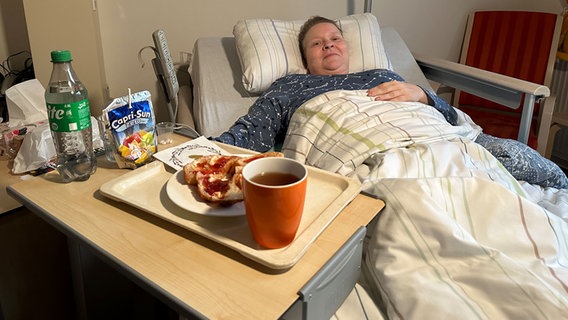 Eine Person liegt in einem Bett und bekommt ein Essen geliefert. © NDR Foto: Johannes Tran