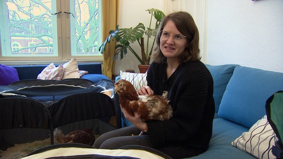 Eine Frau sitzt in ihrem Wohnzimmer mit einem Huhn auf dem Arm. © NDR 