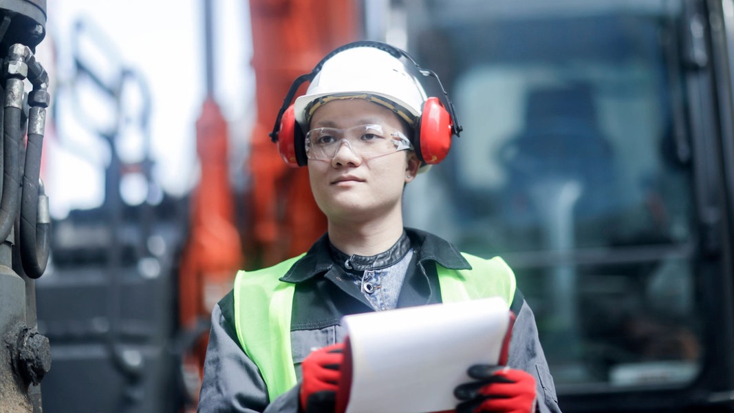Eine Ingenieurin trägt einen Helm und Gehörschutz. Sie steht auf einer Baustelle und hält ein Klemmbrett.