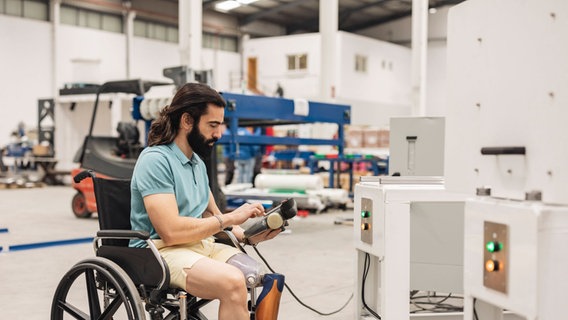 Ein Mann mit Beinprothese sitzt in einem Rollstuhl und bedient eine Maschine in einer Industriehalle. © Imago Images / Westend61 Foto: Westend61