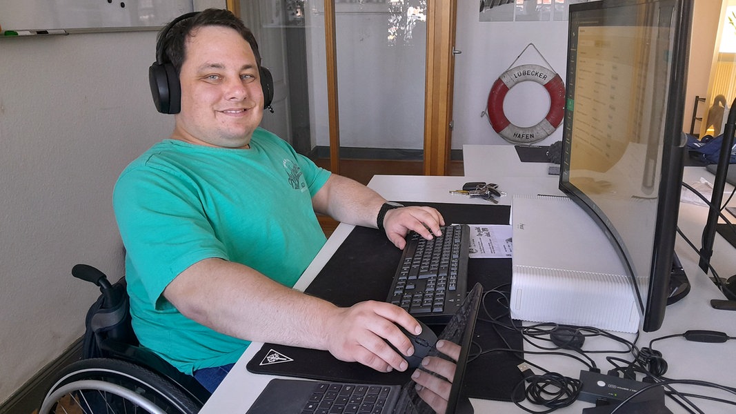 Webdesigner Paul Sznabel sitzt im Rollstuhl vor einem Arbeitstisch mit einem großen Monitor.
