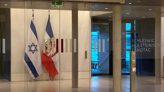 Flagi Izraela i Szlezwiku-Holsztynu stoją obok siebie za szklaną ścianą w parlamencie krajowym.  © NTR Foto: Anna Krusnik
