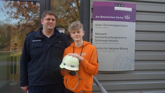 Ein Jugendlicher hält einen Feuerwehrhelm in die Kamera, neben ihm steht sein Vater.  Foto: Stella Kennedy