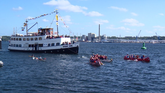 Eine Gruppe von Kanus ziehen ein Fährschiff hinter sich her.  