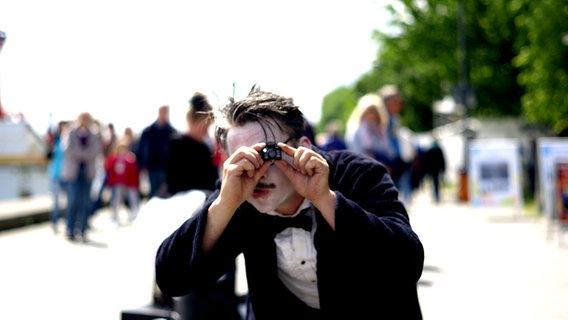 Ein Straßenkünstler hält eine winzige Kamera in der Hand. © NDR Foto: Lisa Pandelaki