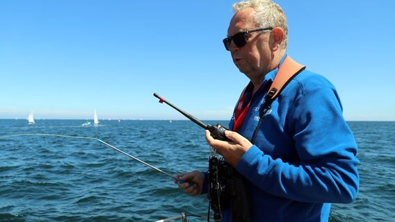 Ein Mann steht an eines Segelschiffs mit einem Funkgerät in der Hand während der Kieler Woche. © NDR Foto: Marina Heller