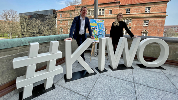 Kiels Oberbürgermeister Ulf Kämpfer (SPD, l.) und eine Frau (r.) stehen hinter großen Buchstaben, die den Hashtag Kieler Woche bilden auf dem Rathaus-Balkon bei einer Pressekonferenz. © NDR Foto: Christopher Gaube
