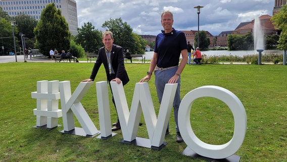 Kiels Oberbürgermeister Ulf Kämpfer (SPD) und Regattaleiter Dirk Ramhorst stehen auf einer Wiese vor weißen Buchstaben "#KIWO". © NDR Foto: Kai Peuckert