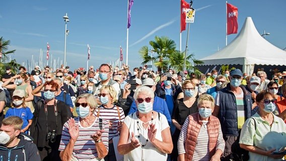 Besucher nehmen mit einem Mund-Nasen-Schutz an der offiziellen Eröffnung der Kieler Woche vor einer Bühne im Olympiastützpunkt Schilksee teil. © dpa-Bildfunk Foto: Gregor Fischer