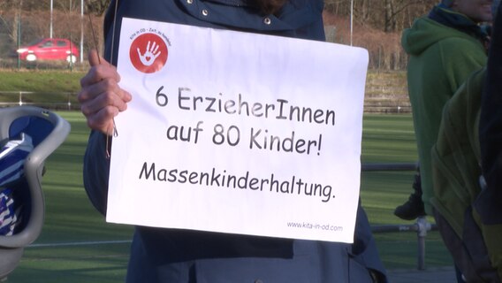 Auf einer Demonstration gegen mangelnde Kitaplätze, hält jemand eine Plakat mit der Aufschrift: "6 ErzieherInnen auf 80 Kinder! Massenkinderhaltung. © NDR 