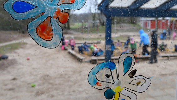 Auf einer Fensterscheibe sind bunte Schmetterlinge gemalt. Dahinter ist unscharf eine Spielfläche mit vielen kleinen Kindern zu erkennen. © dpa-Bildfunk Foto: Carsten Rehder