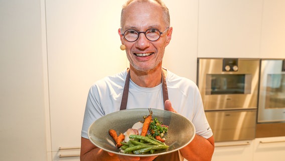 Spitzenkoch Robert Stolz hält lächelnd ein Gericht in die Kamera. © NDR Foto: Julian Marxen