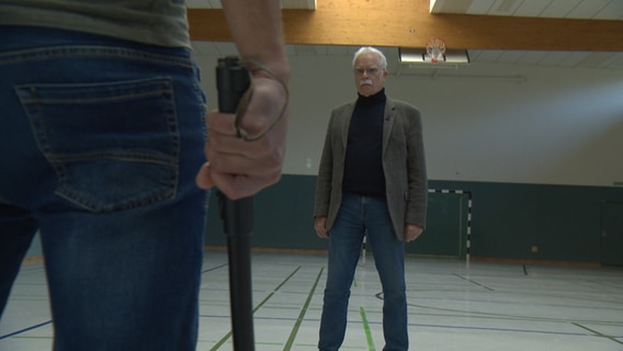 Ein älterer Herr steht in einer Turnhalle, vor ihm steht eine Person mit einem Schlagstock in der Hand.  