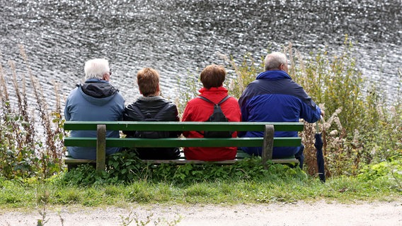Vier Wanderer sitzen auf einer Bank am Ufer eines Gewässers. © IMAGO/blickwinkel 