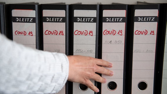 Eine Hand schiebt einen Ordner mit der Aufschrift "COVID-19" zurück in eine Regalreihe. © picture alliance / dpa Foto: Marijan Murat