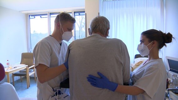 Zwei Pflegende helfen einem Patienten aus einem Stuhl. © NDR 