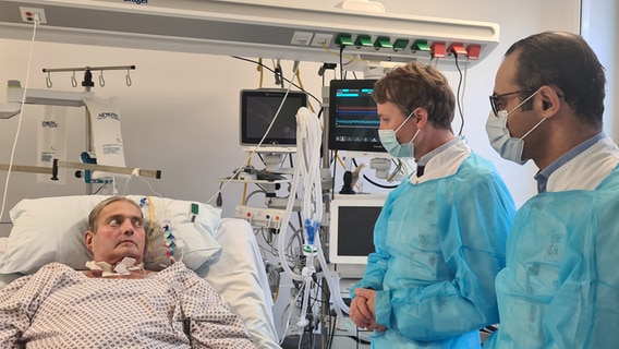 Kunstherz-Empfänger Ralf Jensen liegt im Krankenbett, zwei Ärzte stehen daneben. © NDR Foto: Cassandra Arden