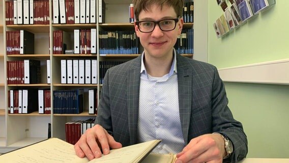 Helge-Fabien Hertz sitzt am Schreibtisch und blättert durch ein Buch. © NDR Foto: Corinna Below