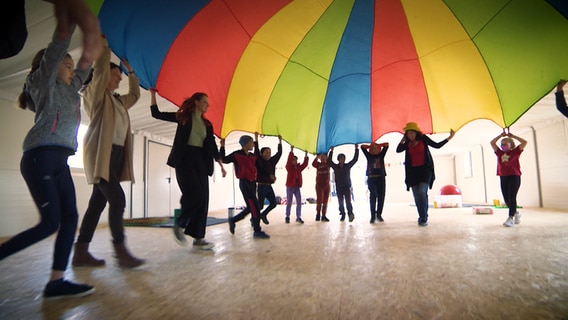 Kinder und Erwachsene laufen im Kreis aufeinander zu und schwingen ein buntes, großes Tuch über sich. © NDR 