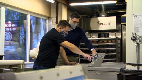 Tino Matthiessen und Jan Pimanow stehen in einem Produktionsraum einer Bäckerei und schauen auf den Bildschirm eines Laptops. Tino Matthiessen und Jan Pimanow stehen in einem Produktionsraum einer Bäckerei und schauen auf den Bildschirm eines Laptops. © NDR