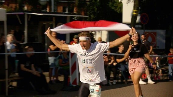 Ein Mann läuft bei einem Triathlon jubelnd und mit einer Fahne in den Händen in die Zielgerade © NDR Foto: NDR