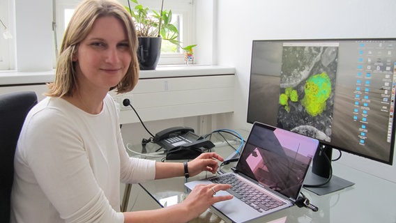 Prof. Dr. Sabrina Jabs ist sitzt in ihrem Büro an der CAU an ihrem Arbeitstisch mit einem Laptop und einem PC-Monitor und lächelt in die Kamera. © Jens Zacharias/NDR Foto: Jens Zacharias