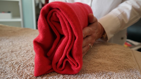 Ein roter Pullover wird von Astrid Saalmann so gefaltet, dass er auf dem Bügelbrett steht. © NDR Foto: Jennifer Lange