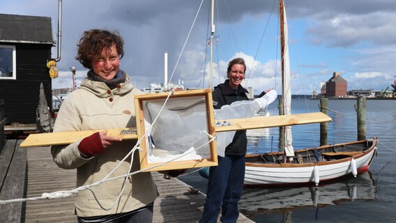 Lauren Grüterich (links) und Caro Höschle (rechts) stehen auf dem Steg des Museumshafens in Flensburg und halten einen selbstgebauten „Manta Trawl“ in die Kamera. © Weniger ist Meer 