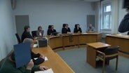 Ein Angeklagter sitzt in einem Gericht und verdeckt sein Gesicht mit einer Mappe. © NDR Foto: NDR Screenshot