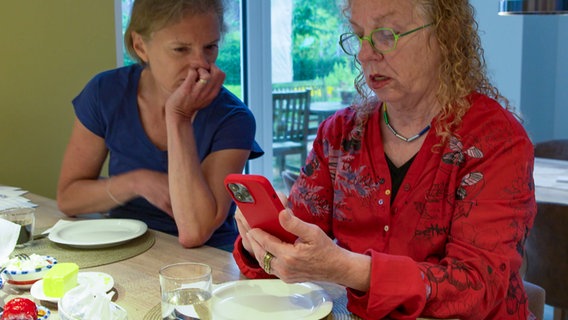 Zwei Frauen sitzen an einem Tisch in einem Wohnzimmer und blicken auf den Bildschirm eines Smartphones. © NDR 