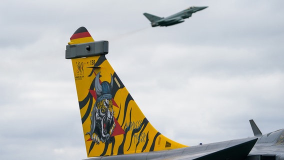 Im Vordergrund ist das Seitenleitwerk eines Kampfjets zu sehen, auf das ein Tiger lackiert wurde. Im Hintergrund fliegt ein Jet in der Luft. © dpa Bildfunk Foto: Marcus Brandt
