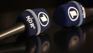 Zwei Mikrofone mit NDR und ARD-Logo liegen auf einem Tisch © dpa/picture-alliance 