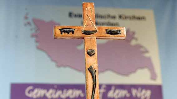 Ein Kreuz steht auf einem Podium vor einem Transparent der Nordkirche. © dpa / Picture Alliance Foto: Carsten Rehder