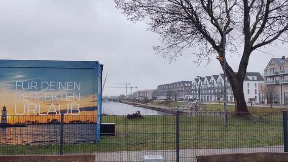 Ein Banner auf einer Gebäuderückseite in Olpenitz hat die Aufschrift "Für Deinen perfekten Urlaub". Rechts sind Hafenbecken und eine Häuserreihe zu sehen. © NDR Foto: Peer-Axel Kroeske