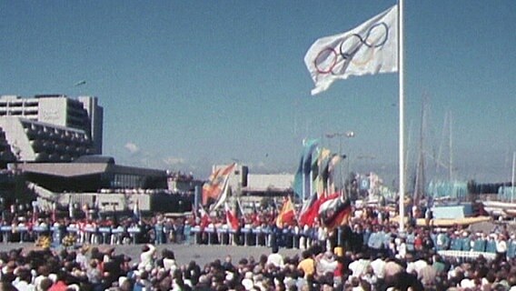 Die olympische Flagge weht über einer Menschenmenge. © ARD 